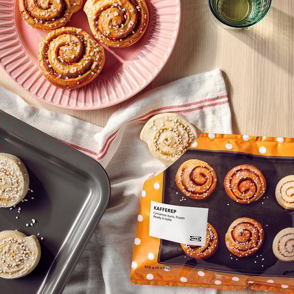 Bilden visar kanelbullar på brickor, vissa bakade och andra i förpackningar märkta KAFFEREP från IKEA, med ett glas vätska på ett träbord.