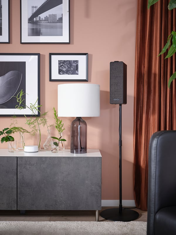 Ett modernt grått sideboard med en bordslampa, växter och en högtalare på stativ, mot en persikofärgad vägg med svartvita bilder.