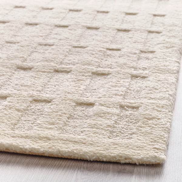 Närbild på en texturerad, off-white matta placerad på ett lätt trägolv. Mattan verkar mjuk och mjuk med ett upprepat mönster av små öglor.