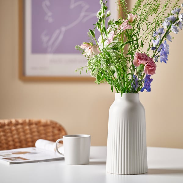 Modern vit, texturerad cylindrisk vas rymmer höga blommor, sitter på bordet bredvid en liten kopp och korg.