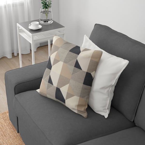 En grå soffa med tjocka kuddar och en mönstrad kudde. Ett sidobord med en växt och koppar som troligen är avsedda för kaffe, vilket tyder på avkoppling och komfort.