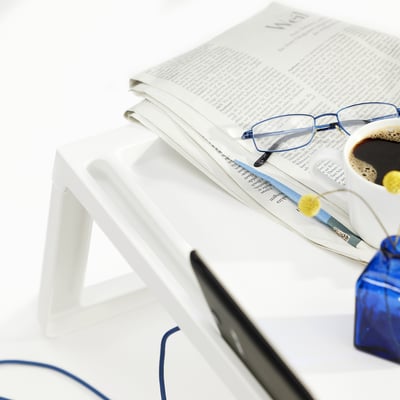 En vit hopfällbar sängbricka rymmer en tidning, glasögon, kaffe och penna, som visas med en delvis synlig surfplatta och vit bakgrund.