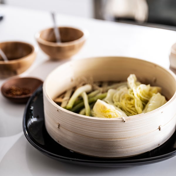 Ångskål av bambu rymmer gröna grönsaker, sitter på svart tallrik med träbehållare.