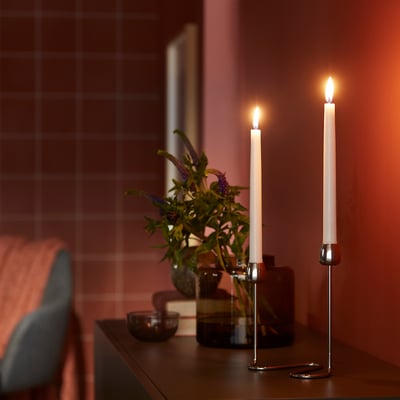 KLOKHET vita ljus i metallhållare; Lång, smal, flimrande på mörk yta bredvid en liten blå vas med blommor, mot den kaklade väggen.