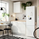 Litet, modernt kök med vita KNOXHULT-skåp, svarta accenter, brun träskiva, svarta stolar och ett högt vitt kylskåp.