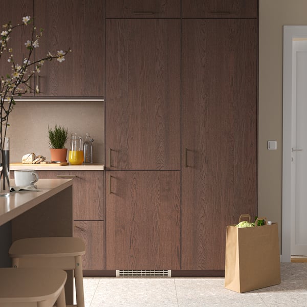 Modernt kök med mörka träskåp, högt KÖLDGRADER-kylskåp, bänkskiva med växter, burkar, bröd och matvaror på golvet, litet barbord.