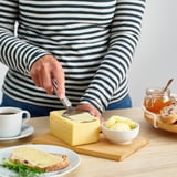 En person skivar ost på en träskiva, med hjälp av en skivare i rostfritt stål. Kaffe, ost på bröd och äpplen finns också.