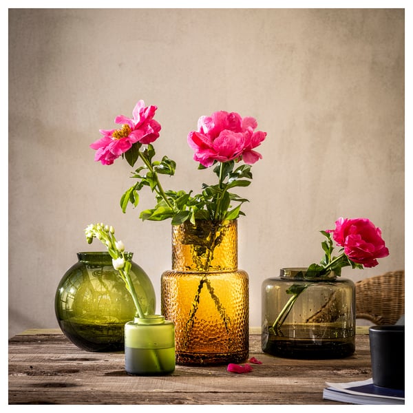 Tre färgglada glasvaser, var och en med blommor inuti, arrangerade på ett rustikt träbord. Design av Ilse Crawford.