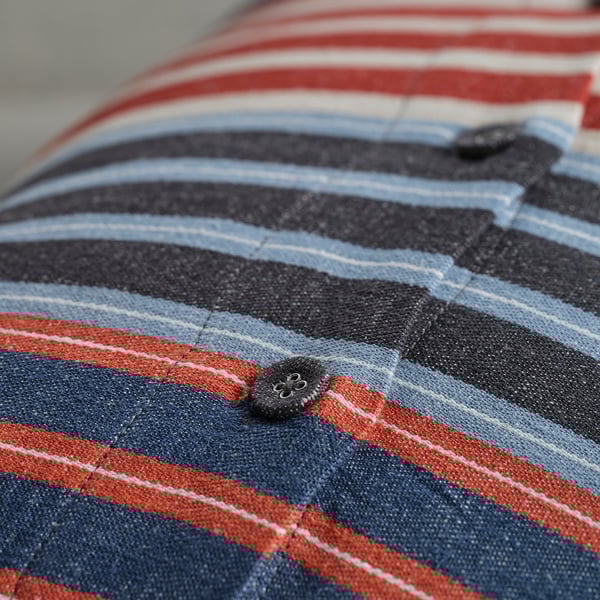 Närbild på randigt kuddfodral med tygklädd knapp. Mörkblå, röda och vita ränder på texturerat tyg.