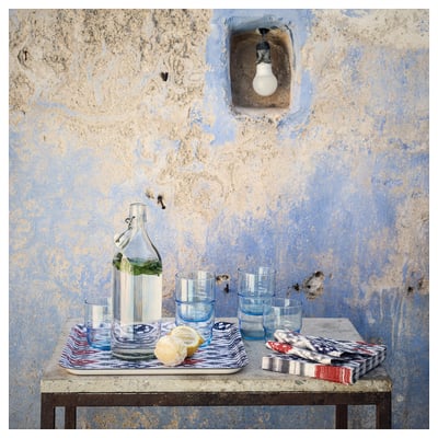 Klar KORKEN glasflaska med citron och glas. Väderbiten blå vägg och glödlampa. Mönstrade textilier på bordet.