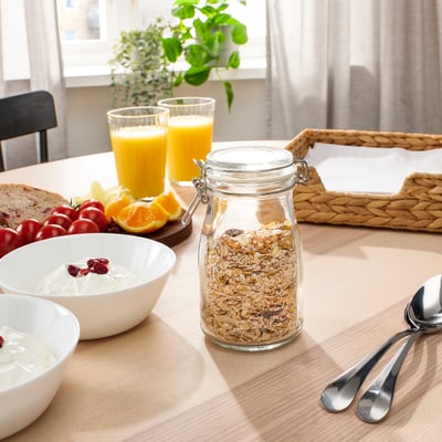 Klar glasburk med granola, förseglad med metallklämma, sitter på ett träbord. Skål med yoghurt med tranbär, apelsinjuice och sked bredvid den.