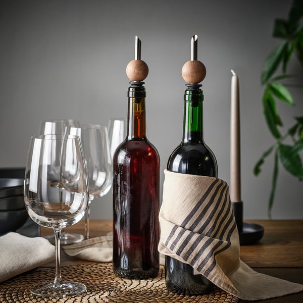 Två vinflaskor med droppproppar i trä på mörkt bord, bredvid vinglas och en trasa.