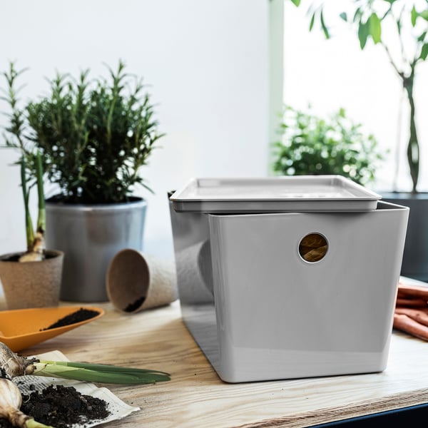 Grå KUGGIS låda med handtag, omgiven av växter och verktyg. Medelstor, tillverkad av återvunnen plast.