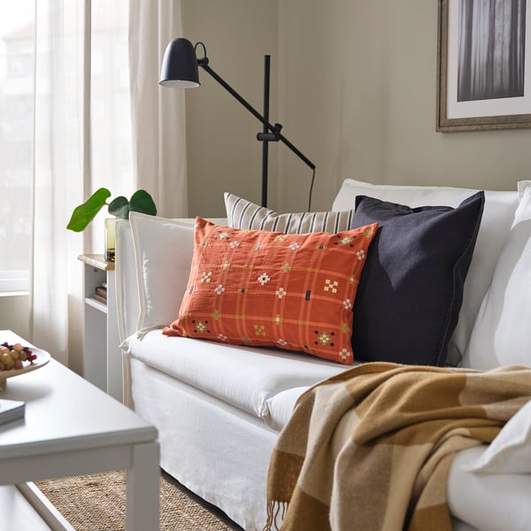 Ljust vardagsrum med KUSTGRAN orange kuddar på en vit soffa. Svart golvlampa bredvid, beige gardin i bakgrunden.