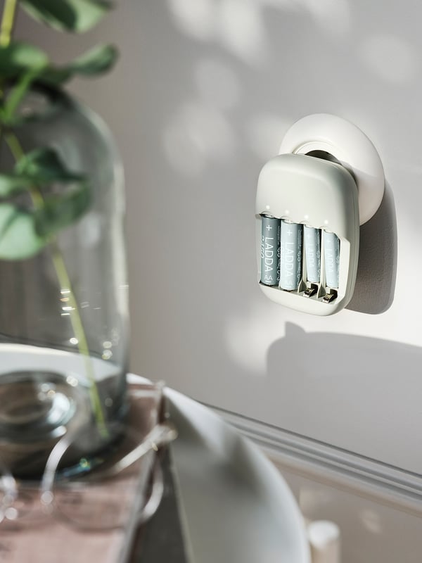 En väggmonterad vit LADDA batteriladdare med tre silverfärgade uppladdningsbara batterier inuti. En del av en grön växt syns bredvid den.