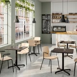 Modernt café med träbord och beige stolar. Hängande växter, stora fönster, svarta lampor och en svart tavelmeny skapar en avslappnad atmosfär.