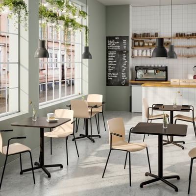 Modernt café med träbord och beige stolar. Hängande växter, stora fönster, svarta lampor och en svart tavelmeny skapar en avslappnad atmosfär.