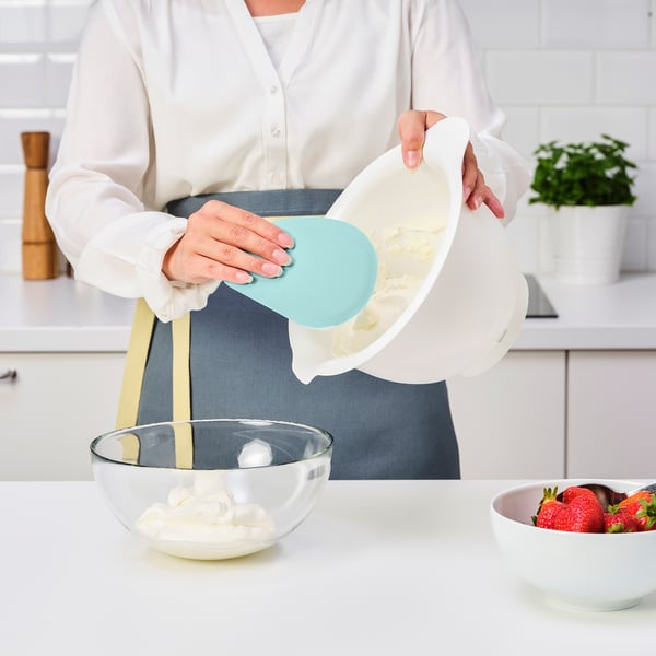 Person som häller smet från en vit skål med hjälp av en blå spatel i en glasskål på en köksbänk, bredvid jordgubbar.