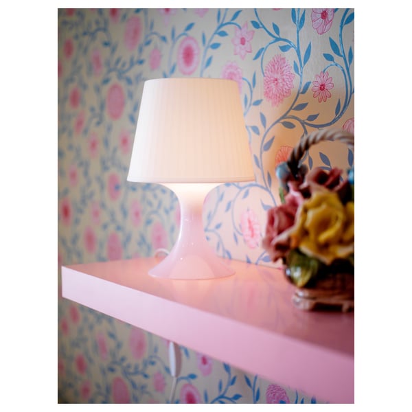 En vit lampa, LAMPAN, står på en rosa yta och avger ett mjukt varmt ljus. Dess enkla design och klotformade glödlampa skapar en mysig atmosfär.