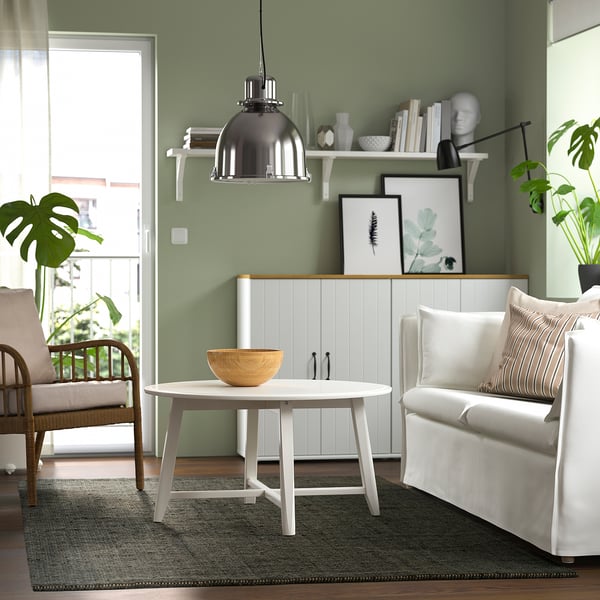 Modernt vardagsrum med LANDBANA jutematta. Mörkgrön, rektangulär matta under ett vitt bord. Korgstol och beige soffa på matta. Grön fondvägg.
