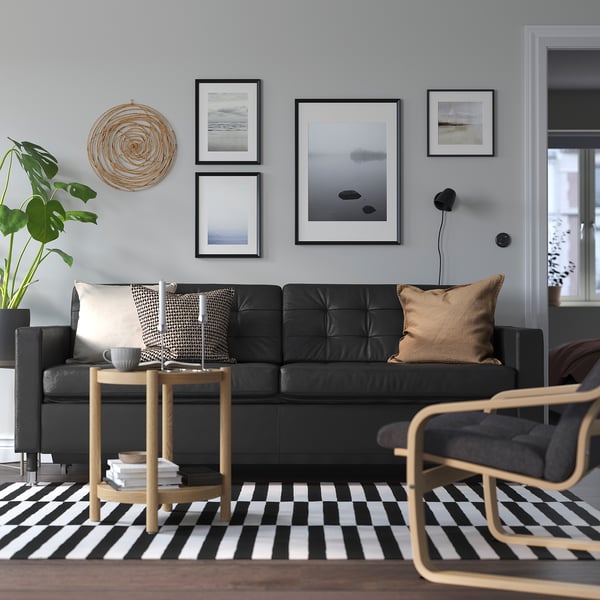 Modernt vardagsrum med svart skinnsoffa, konst, matta, sidobord, växt och lampa.