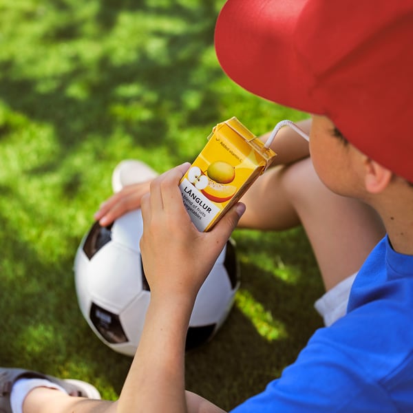 Barn dricker gul lang lur smoothie, med fotboll, bär röd keps.