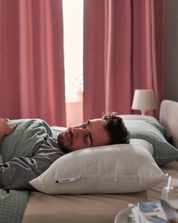 En man sover på en hög vit LAPPTÅTEL-kudde, vars mikrofibertyg är mjukt och stödjande. Bredvid honom ramar ett rött draperi in ett fönster.