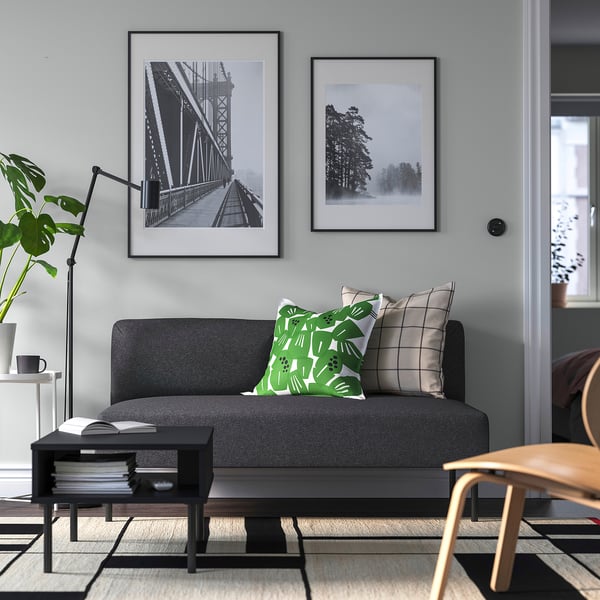 Modernt vardagsrum med grå LILLEHEM soffa, gröna kuddar, svart soffbord, trästol, matta, lampa, böcker, konst.