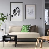 Modernt vardagsrum med lillem beige soffa, svart soffbord, två inramade konstverk och växter. Soffkuddar i grönt och svart.