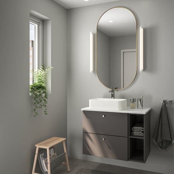 Modernt badrum med stor guldinramad spegel, mörkgrå svävande kommod, träpall, växt och grå handdukar.