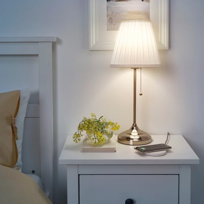 Nattduksbord med tänd LIVBOJ-lampa, bok och blomvas; Ljusblå sänggavel, beige kudde, vita väggar.