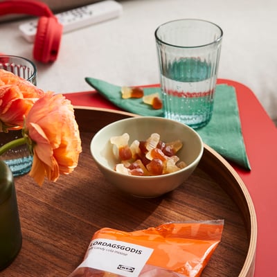 Skål med svenska gelégodis med colasmak i björnform på en bricka med ett glas vatten, blomma och godisförpackning.