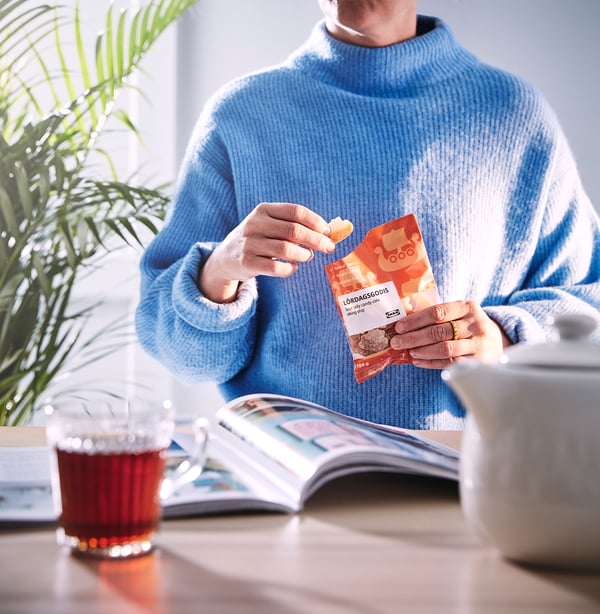 Person i blå tröja som rymmer LÖRDAGSGODIS godis, sittande vid bordet med te, tidskrift och växt.