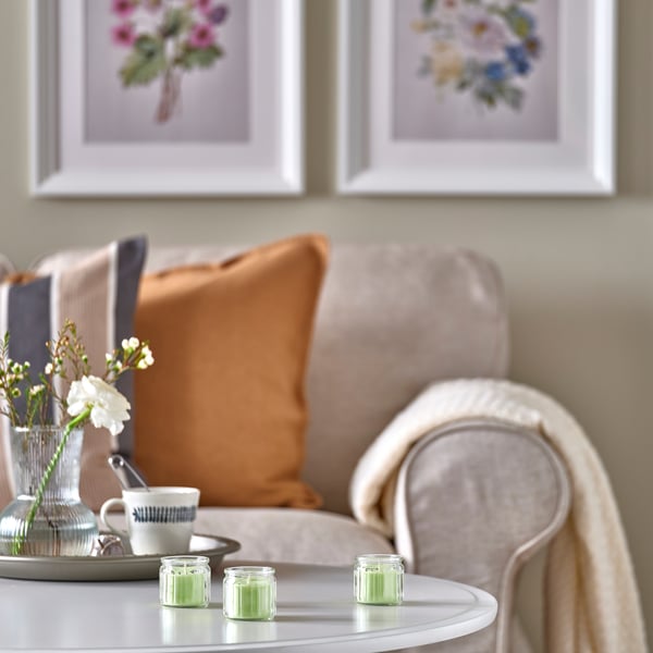 Tre gröna ljus på bordet nära soffan med kuddar, blommor och tryck.