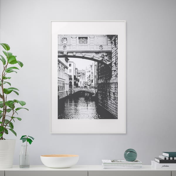 Inramad kanalscen med bro, hyllhållande växt, vas, skål, böcker, sfär.