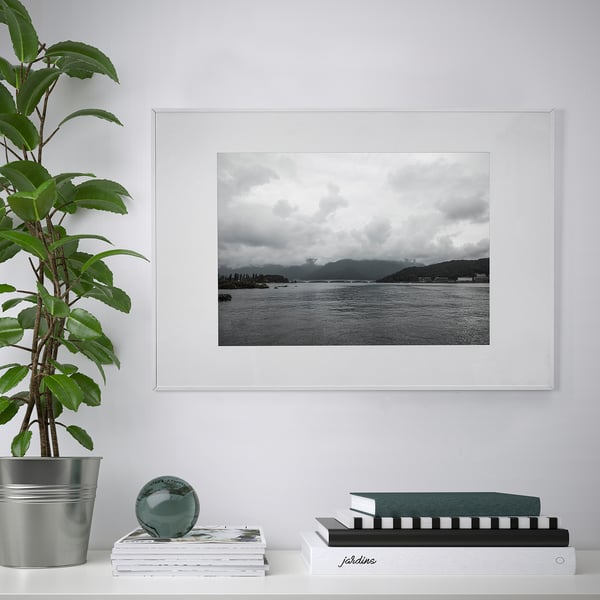 Vit LOMVIKEN fotoram med svartvit maritim bild, placerad ovanför böcker och ett dekorativt föremål. Grön växt till vänster.