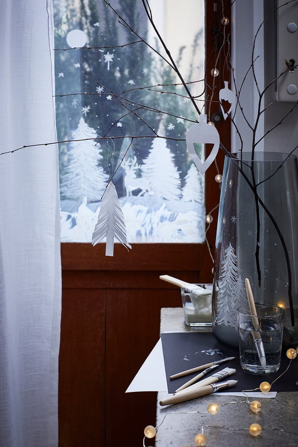 Snöigt fönster med vas, kala grenar och vita pappersprydnader, mjukt upplyst.