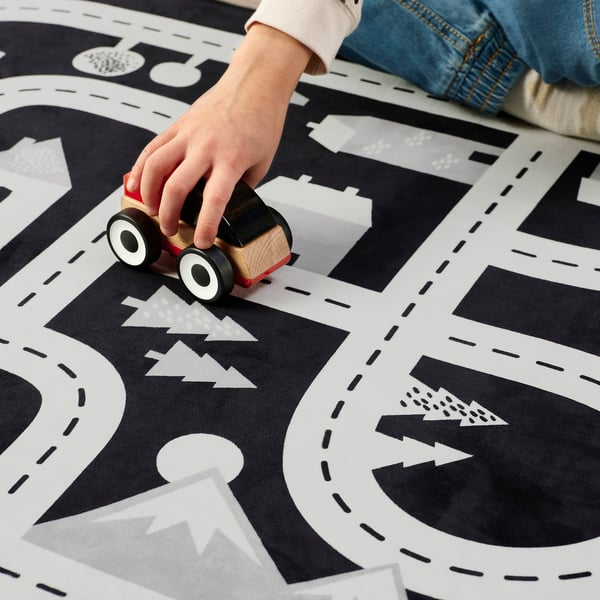 Barnens handstyrning av träbil på svart och vit lekmatta med stads- och skogsdesign.