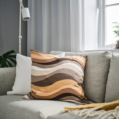 Modernt vardagsrum med kudden MYGGBLOMSTER, bruna toner, på grå soffa, nära krukväxt, vit lampa, fönster med skira gardiner.