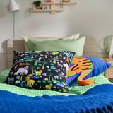 Färgglada kuddar, grönt täcke, vit sänggavel, växt, lampa, hyllor.