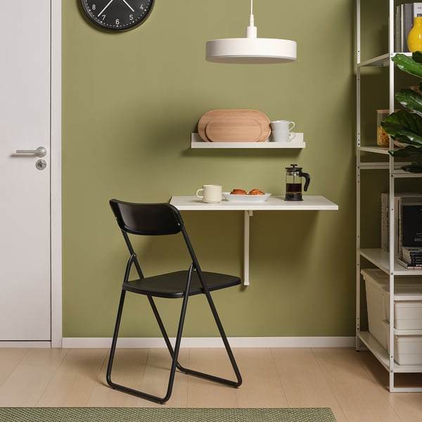 Litet, väggmonterat vitt bord, med svart fällstol. Modern design, grön vägg, hängande klocka och växt.