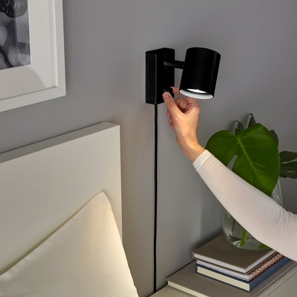 En person justerar en väggmonterad NYMÅNE-lampa bredvid en säng och bokhylla med en växt.