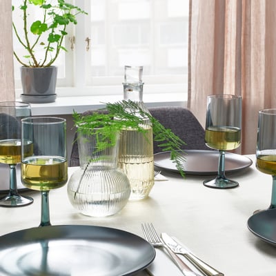 Bordsuppsättning med OMBONAD blå tallrikar, klara texturerade glasvaser som håller grönska och vinglas på vit duk nära fönstret.