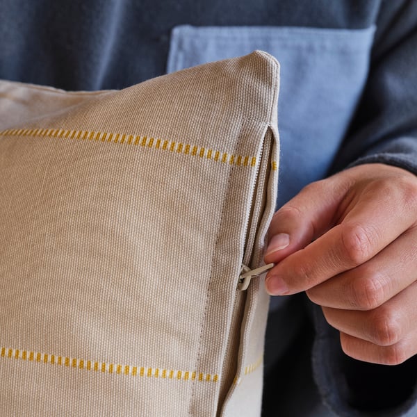Närbild av en hand som håller ett beige kuddfodral i bomull med gula geometriska mönster. Dragkedjan är delvis öppen, vilket visar att den är lätt att använda.