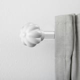 Vit keramisk gardinfinial som liknar en blomknopp, säkert fäst vid en grå gardin på en vit vägg.