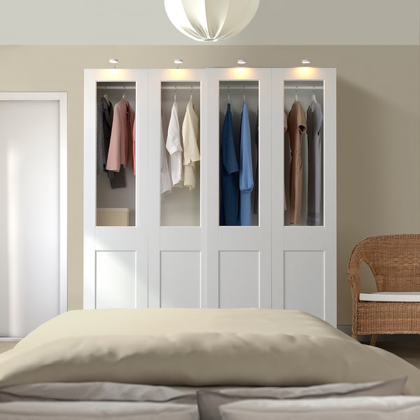 Stor vit PAX garderob med glasdörrar och kläder. Säng med beige sängkläder fram, korgstol i närheten.