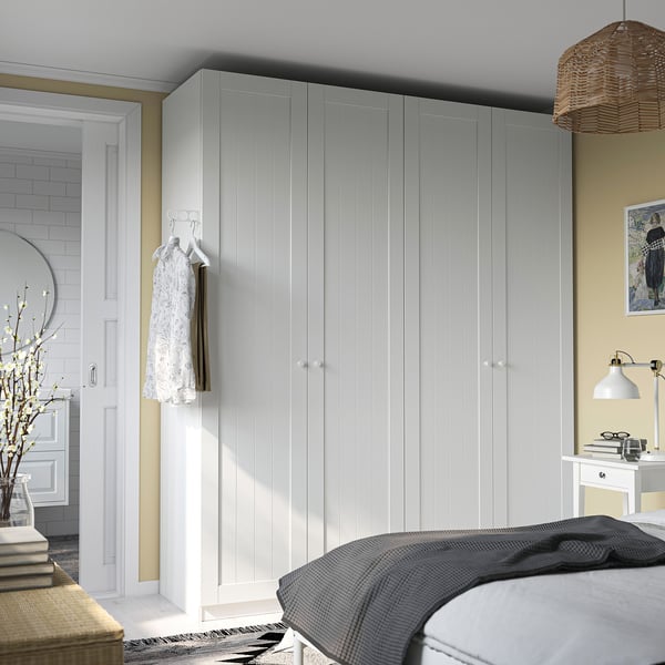 Den stora vita PAX-garderoben sträcker sig över rumshöjd och rymmer hängande plagg, sett med ett beige sovrum med en säng och sidobord.
