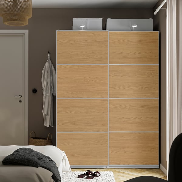 En brun IKEA PAX-garderob med skjutdörrar, högre än säng och dörr, ekmönstrad folie, bredvid ett fönster med beige gardiner.