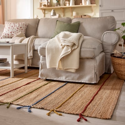Handvävd jutematta med färgglada ränder och ulltofsar i vardagsrummet, under beige soffa och sidobord.