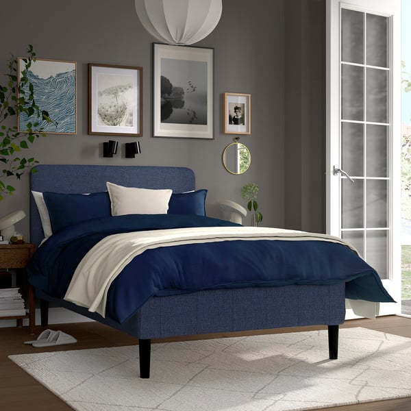 Modernt sovrum med blå queen size-säng, marinblå sängkläder, grå väggar, inramade bilder, frostad glasdörr, matta, spegel, nattduksbord med lampa och växt.